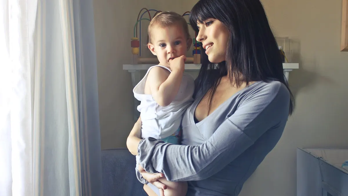 Une mère souriante tient contre elle son bébé, qui regarde l'objectif, près d'une fenêtre lumineuse