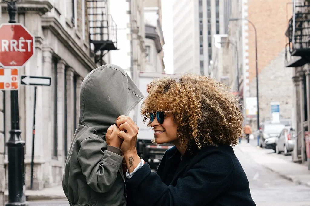 Dans la rue, une mère accroupie tient les mains de son enfant, le visage caché sous sa capuche comme pour un jeu de coucou