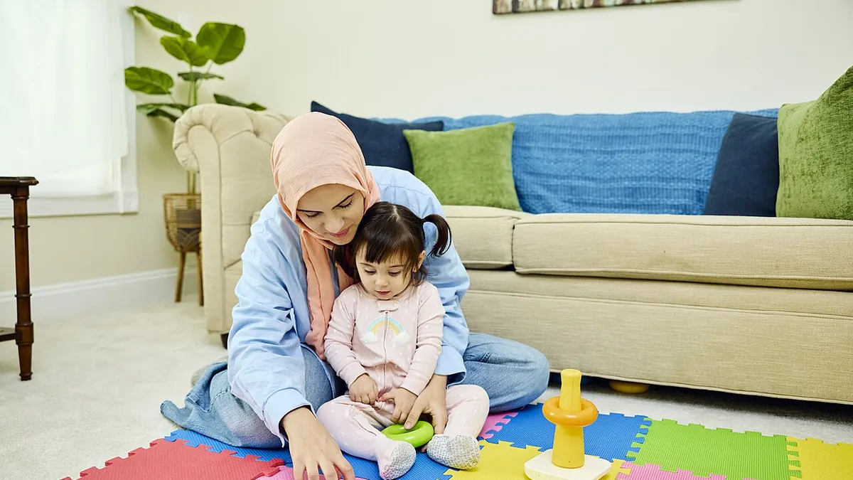 Une femme assise par terre joue avec une bambine sur des tapis en mousse colorés, dans un salon