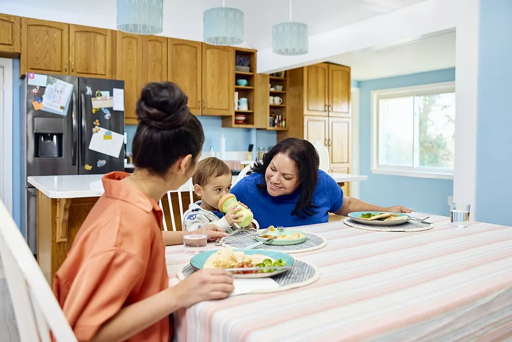 Autour d'une table de cuisine, deux femmes partagent un repas avec un bambin qui boit à son gobelet