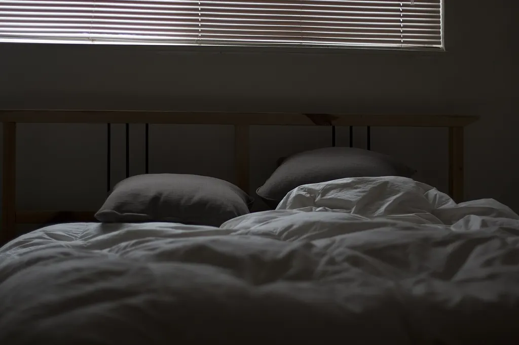Un lit défait dans une chambre sombre, les stores baissés laissant filtrer un peu de jour