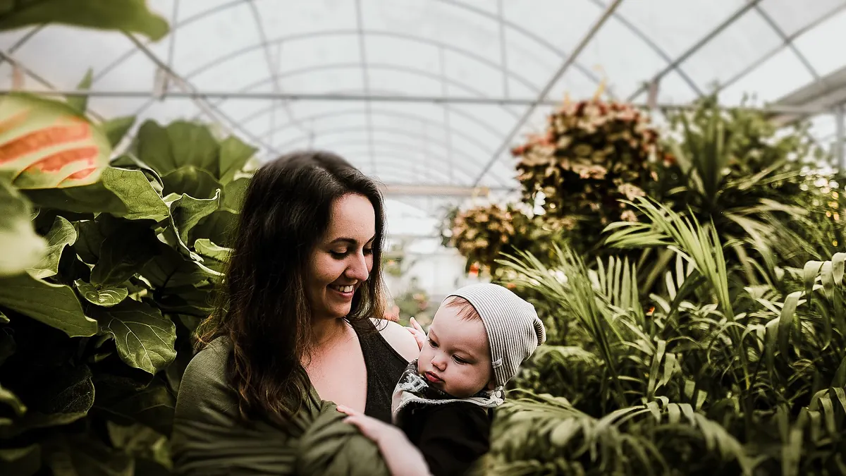 Une mère souriante tient dans ses bras son bébé éveillé, coiffé d'un bonnet rayé, dans une serre