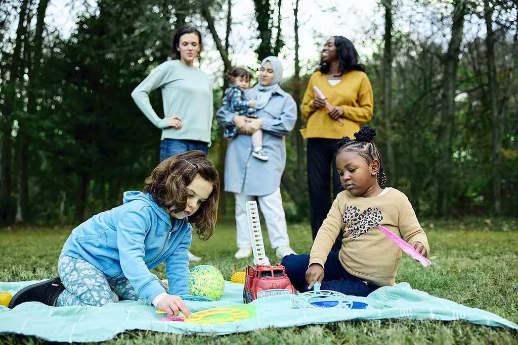 Dans un parc, des parents discutent debout pendant que de jeunes enfants jouent sur une couverture étalée dans l'herbe