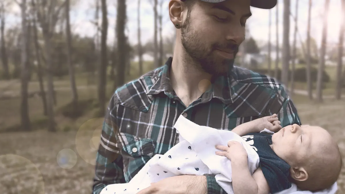 Un père en casquette porte son bébé dans les bras et le regarde, en pleine nature