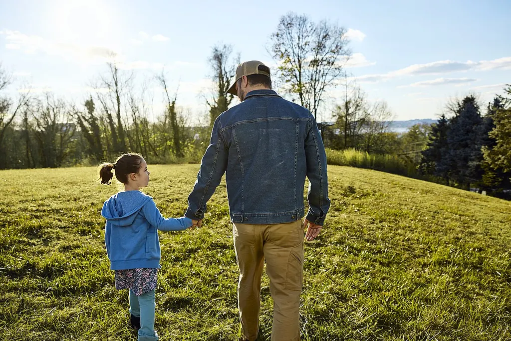 Un père et sa petite fille marchent main dans la main dans un grand pré ensoleillé, vus de dos