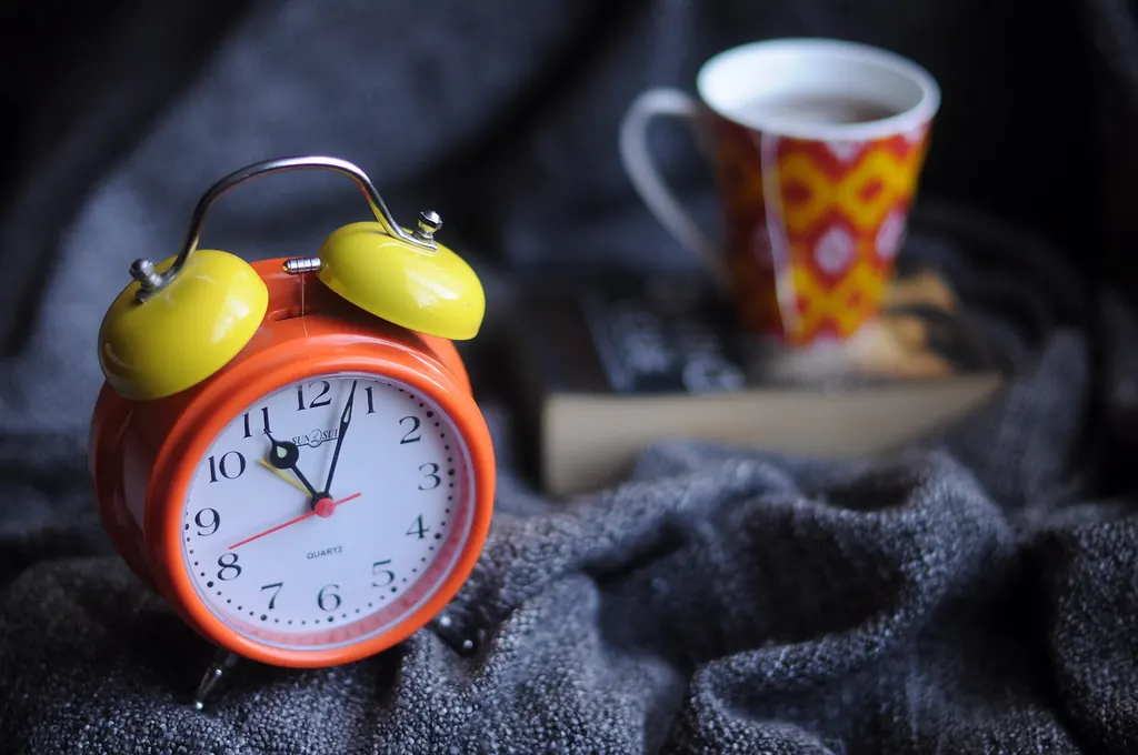 Un réveil orange à côté d'une tasse de café et d'un livre, posés sur une couverture grise