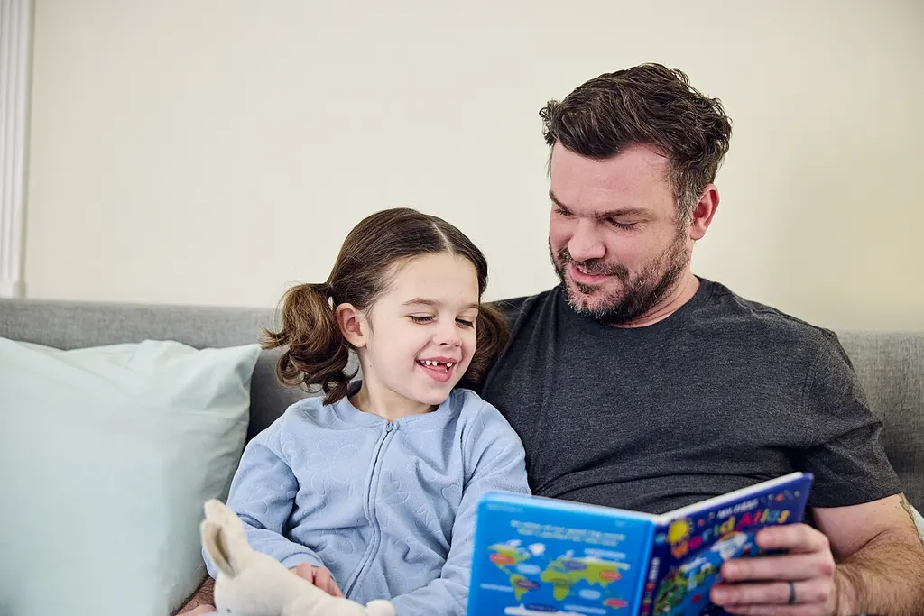 Un père lit un livre à sa fille en pyjama, une peluche contre elle, tous deux assis sur le canapé
